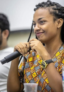 Mulher negra de tranças, de perfil sorri com enquanto segura um microfone com duas mãos. Tem um relógio preto na mao esquerda.