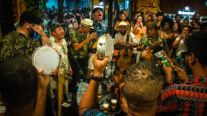 foto de uma roda de samba, onde alguns homens portam instrumentos de percussão, violão e cavaquinho e algumas pessoas em volta fazem registro com o celular