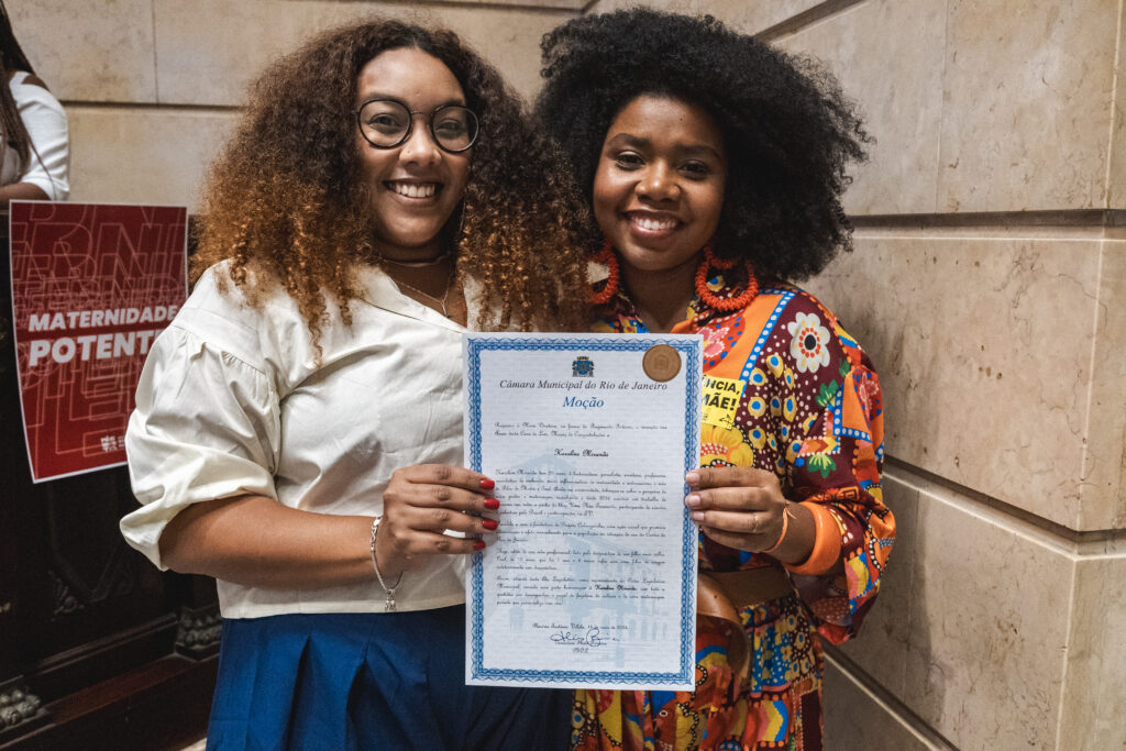 Duas mulheres negras, uma vestindo um vestido estampado e outra uma blusa branca com calça azul marinho, posam para a foto segurando um documento de Moção de Louvor