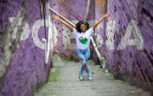 Mulher jovem negra, de óculos, camisa branca estampada, calça jeans e tênis de braços abertos. Ela está posando em um dos degraus de uma escada da periferia, com paredes de cor lilás ao lado. No fundo, está a palavra POESIA