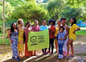 Mulheres negras reunidas enquanto seguram uma bandeira amarela com os dizeres ˜Vozes negras pelo clima"escrito em preto, com a imagem de um punho cerrado compondo um cabelo black feminino. Ao fundo, a paisagem é composta por árvores.