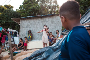 Adolescentes reunidos em uma espécie de laje. em primeiro plano, um menino observa uma menina, ao centro, tocando violino