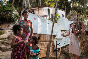 em uma favela, uma mulher negra e duas crianças estão ao lado de uma menina tocando violino