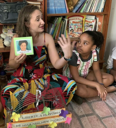 Mulher branca falando sentada no chão com um livro na mão ao lado de uma criança preta