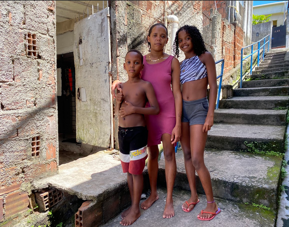 Foto em frente a uma escadaria na favela, em que há uma mulher negra magra vestida de rosa, uma adolescente negra ao seu lado e um menino negro do outro