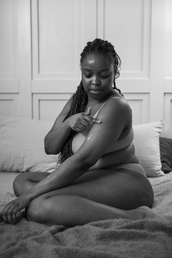 foto em preto e branco com uma mulher negra apenas de calcinha e sutiã está sentada na cama passando creme em seu braço