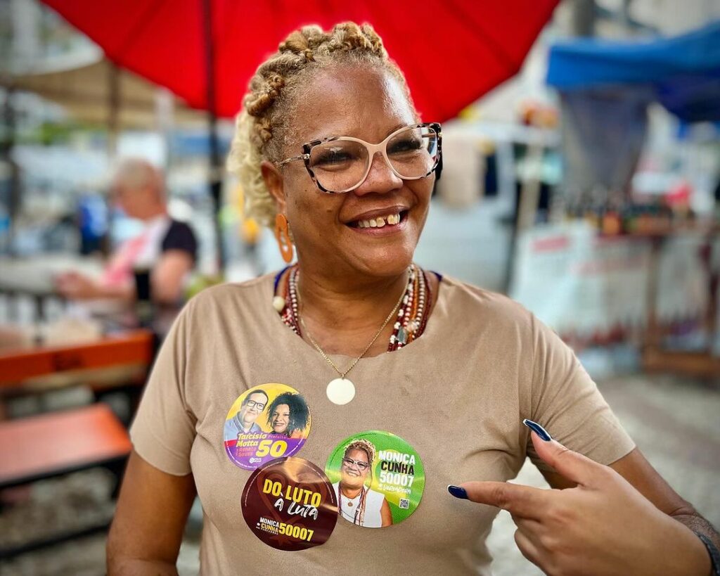 mulher negra sorrindo aponta para adesivo colado na blusa