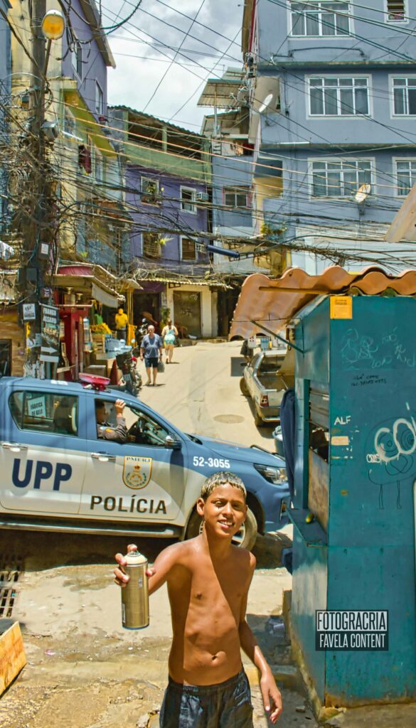 Breninho aparece sorrindo e segura uma lata de tinta spray; atrás dele há um carro da Unidade de Polícia Pacificadora (UPP) e casas da favela.