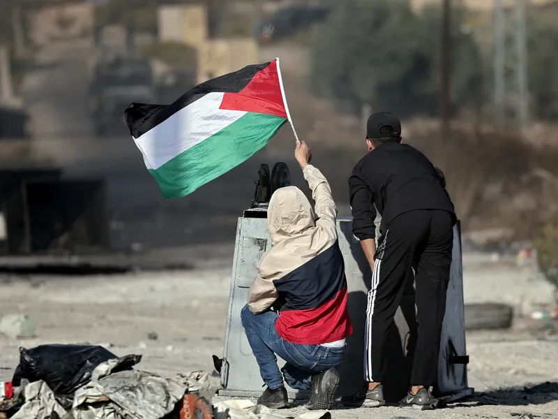 duas pessoas de costas para a foto e de frente para um bombardeio. uma delas levanta a bandeira da Palestina