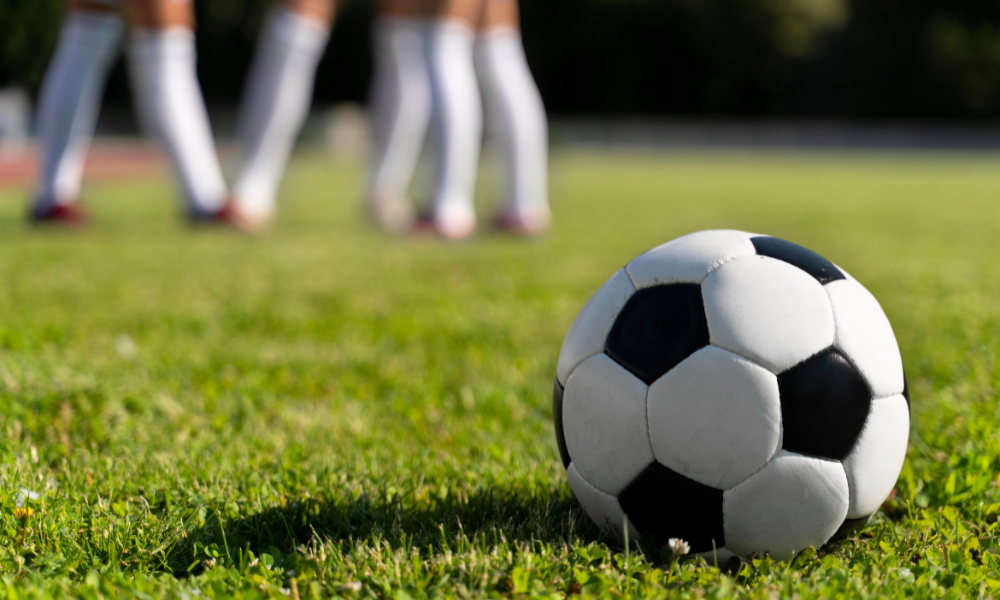 Foto mostra bola em campo de futebol e jogadores ao fundo
