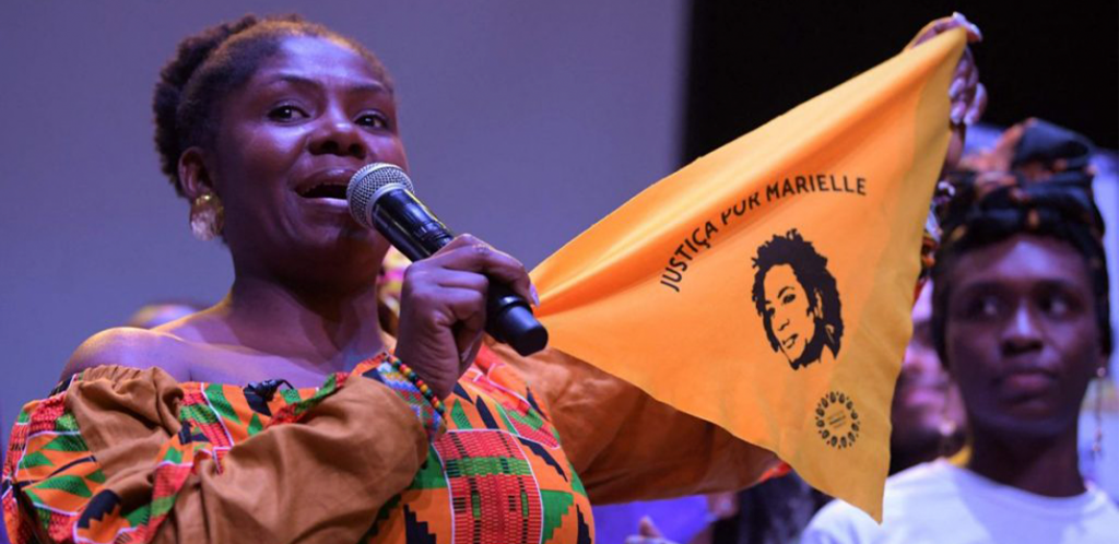 Foto mostra Francia, uma mulher negra, segurando uma bandeira com o rosto de Marielle Franco
