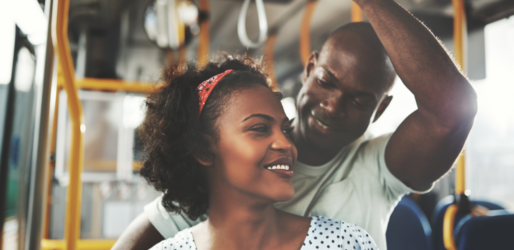 Foto mostra um homem e uma mulher, os dois negros, sorrindo dentro de um ônibus