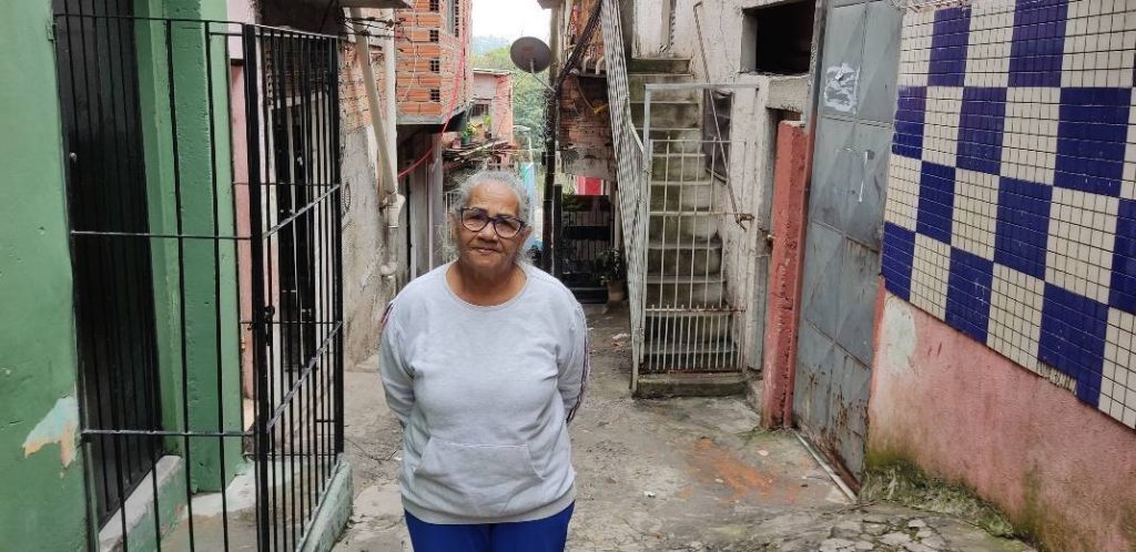Mulher idosa, usando calça azul e blusa cinza, na viela de uma favela