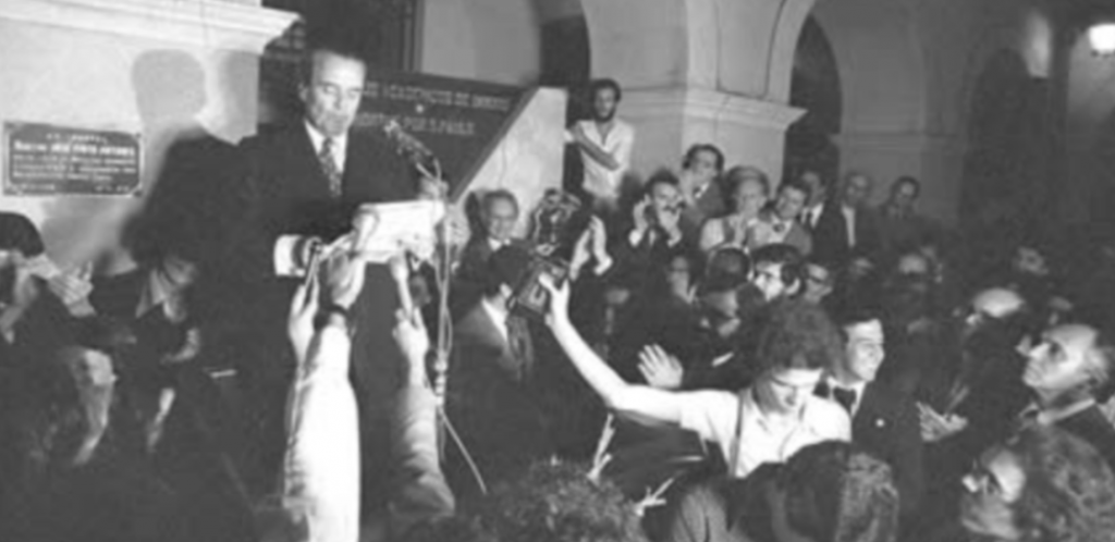 Foto em preto e branco mostra homem branco lendo carta, ao ser redor várias pessoa com microfones.