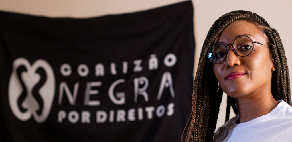 Foto mostra Maíra Vida séria em frente à bandeira da Coalizão Negra por Direitos