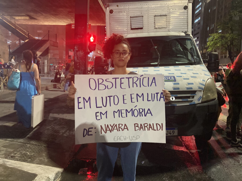 Foto mostra mulher com o cartaz "obstetrícia em luto e em luta em memória de Nayara Baraldo"