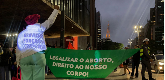 Foto mostra mulher com o punho erguido. Em seu caso, reflete o texto gravidez forçada é tortura. A sua frente, bandeira verde com os dizeres legalizar o aborto / direito ao nosso corpo!