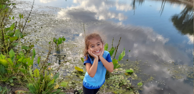 Foto mostra criança sorrindo e, ao fundo, lago com vegetações