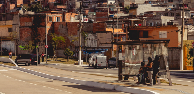Foto mostra pessoas esperando ônibus e, ao fundo, favela