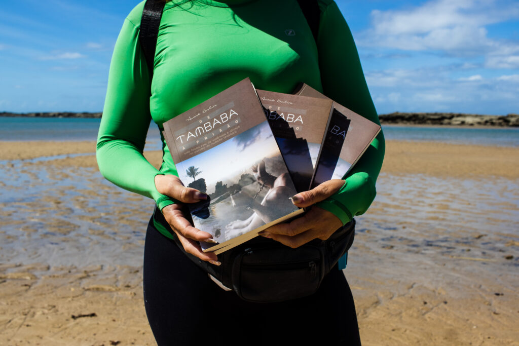 Foto mostra Amanda Karoline segurando seus livros. Ao fundo, o mar da praia de Tambaba