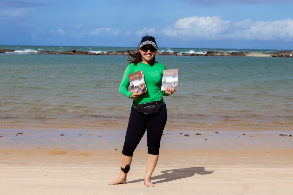 Foto mostra Amanda Karoline segurando seus livros. Em uma das pernas, Amanda tem uma tornozeleira eletrônica. Ao fundo, o mar da praia de Tambaba