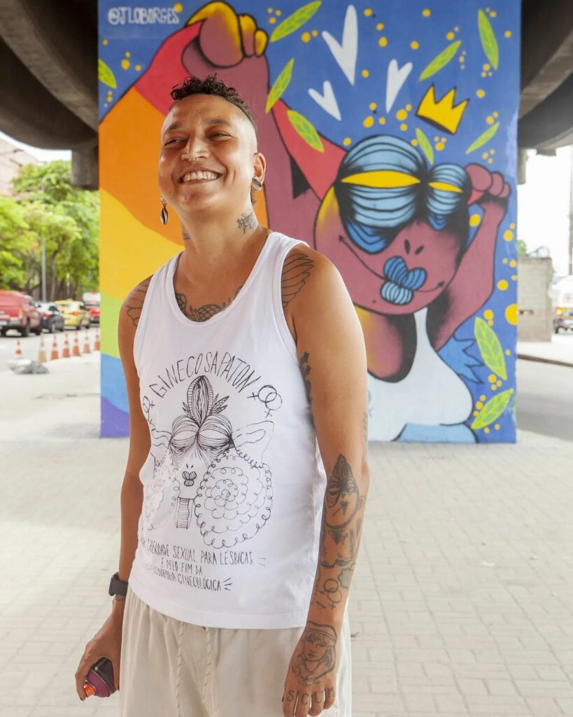 Foto mostra a grafiteira J Lo Borges e ao fundo sua obra